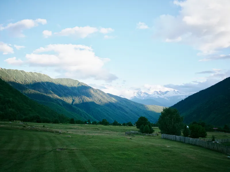 Valley and mountain landscape with green fields, scattered trees, and distant snow capped peaks under a bright blue sky, exuding tranquil nature for outdoor, travel, and countryside themes