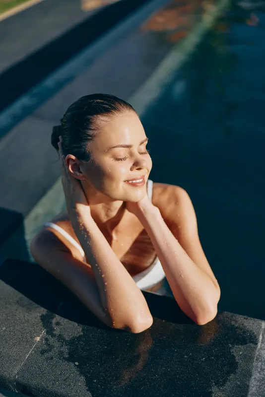 Relaxed woman at pool edge with eyes closed, enjoying sun, peaceful outdoor moment, casual swimwear, warm light, calm mood and self care, by a modern pool with reflective water