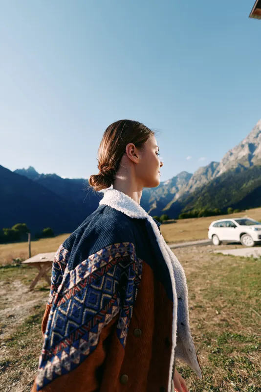 Woman profile in mountains countryside wearing patterned shearling coat, standing in grassy field near parked car. Outdoor portrait, scenic landscape, travel and autumn mood.