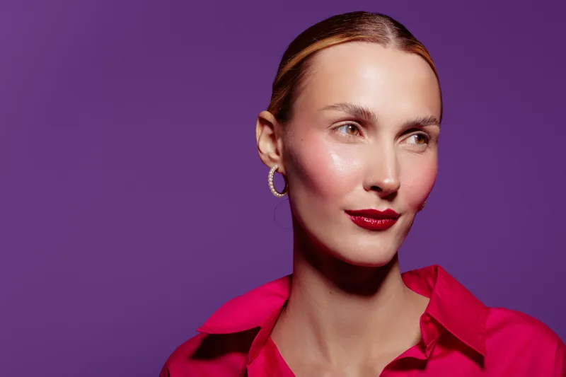 Portrait of a confident woman with slicked back hair wearing red lipstick and a bright pink shirt against a purple background. Concept of fashion, beauty, and elegance in studio lighting.