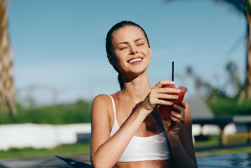 A young woman in a white swimsuit enjoys a refreshing drink outdoors by the pool on a sunny day. She smiles with eyes closed, embodying summer lifestyle and relaxation.