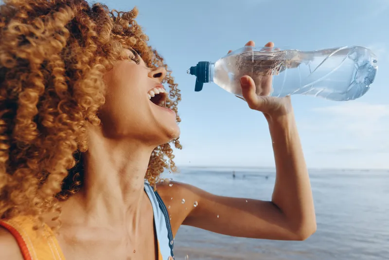 Young woman with curly hair drinking water on a sunny beach, outdoors, in activewear. Focus on hydration, fitness, wellness, outdoor activity and a carefree summer mood.