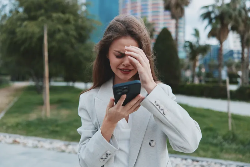 upset woman in gray blazer looking at smartphone with stressed expression outdoors in urban park, financial crisis concept