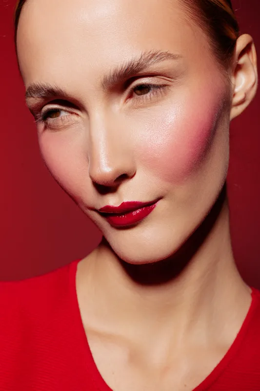 Close-up of a woman with red lipstick and pink blush on cheeks wearing a red top against a red background. Concept of beauty, makeup, fashion, and portrait photography.