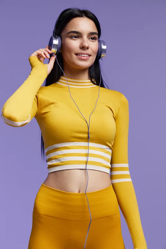 Sport fitness woman wearing yellow activewear and headphones, smiling confidently against purple background, showcasing energy and motivation for healthy lifestyle.
