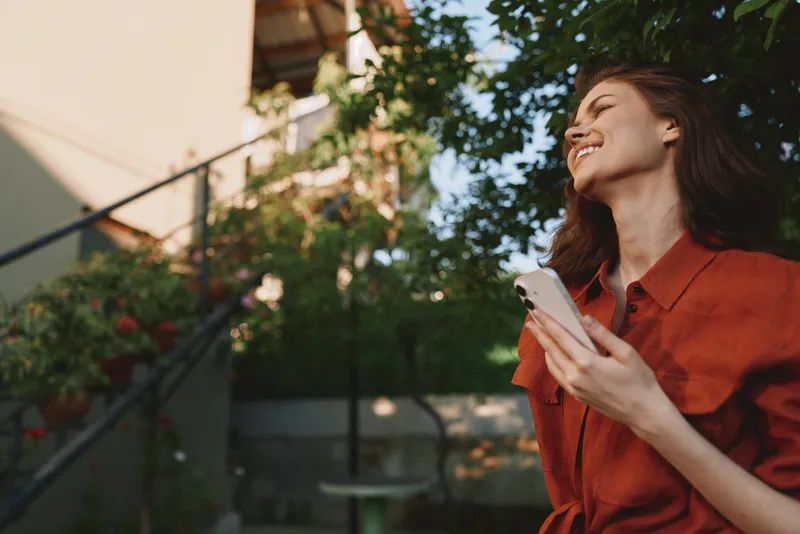 A joyful woman in a red shirt stands outdoors, smiling while scrolling on a smartphone, capturing a casual lifestyle moment in warm daylight, surrounded by greenery and soft light