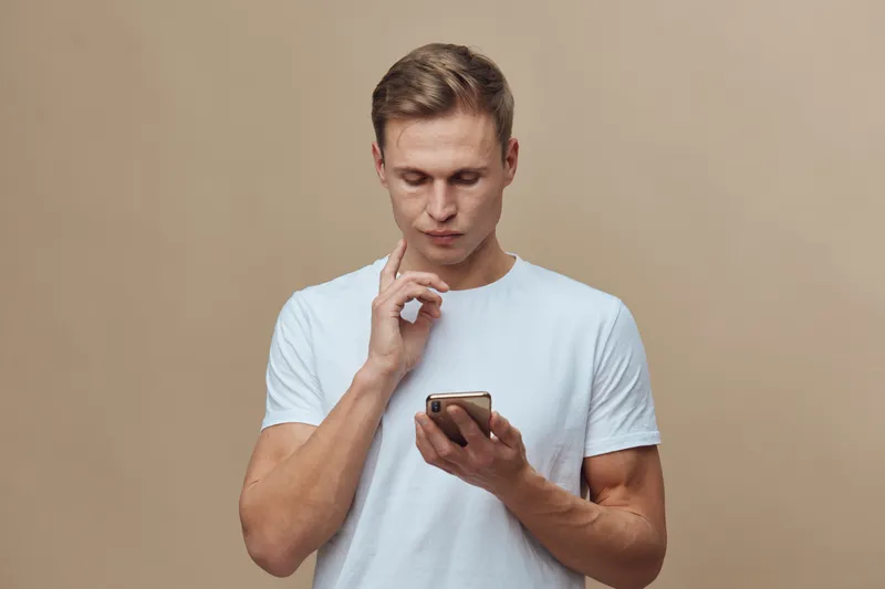 Young caucasian man focused use smartphone mobile phone with serious face wearing white t-shirt isolated beige background digital nomad concept