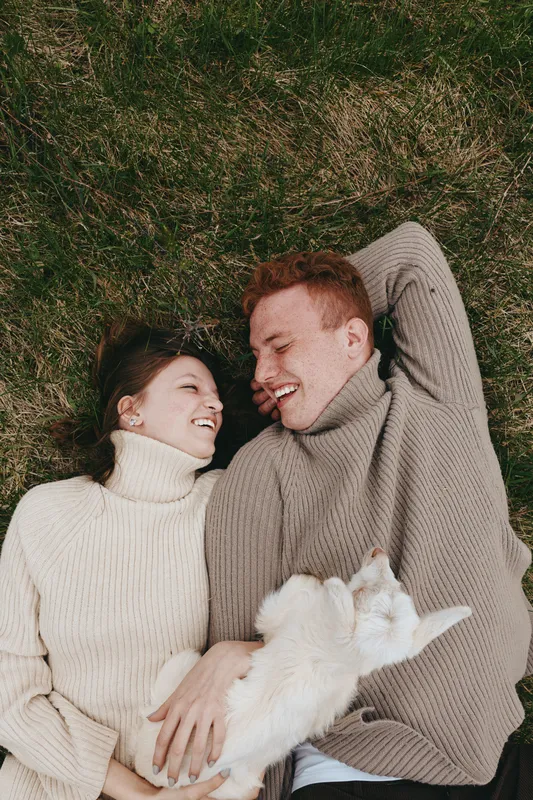 Candid couple lying on grass in cozy sweaters, sharing affection with a playful pet dog, enjoying a relaxed outdoor moment, and warm connection in natural light and gentle sunlight glow