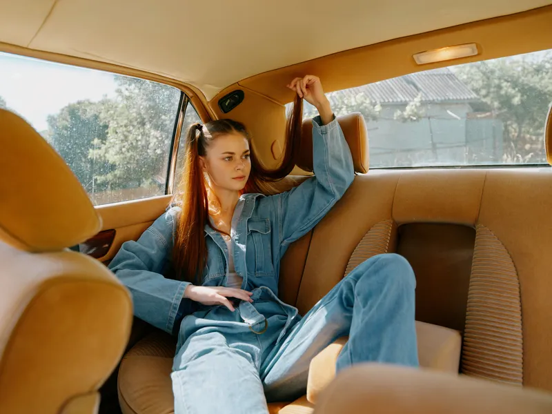 Young woman in denim outfit relaxing in the backseat of a car, enjoying a sunny day, showcasing comfort and casual style, with a natural background.