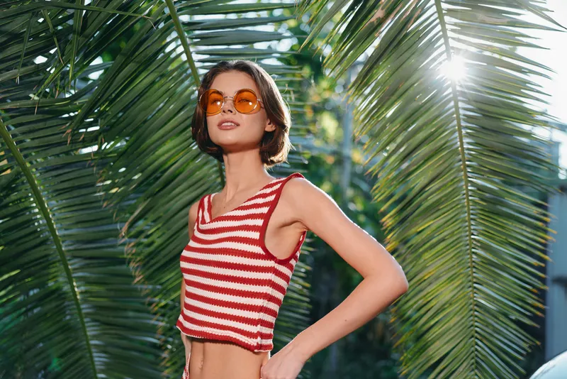 A stylish woman wearing a red and white striped crop top and orange sunglasses stands among tropical palm leaves, posing confidently in sunny outdoor scenery.