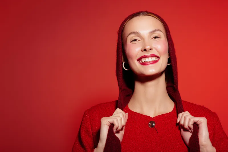 Smiling woman in red sweater and hood poses against red background. She shows happiness and confidence with bright lipstick and hoop earrings. Concept of fashion and joy.