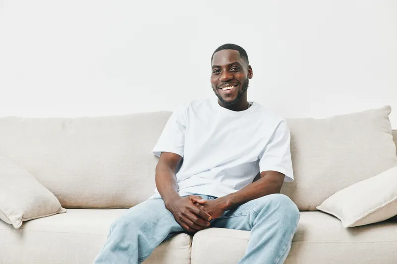 Smiling black man in casual attire sitting on a light colored couch, conveying happiness and comfort in a cozy, bright living space