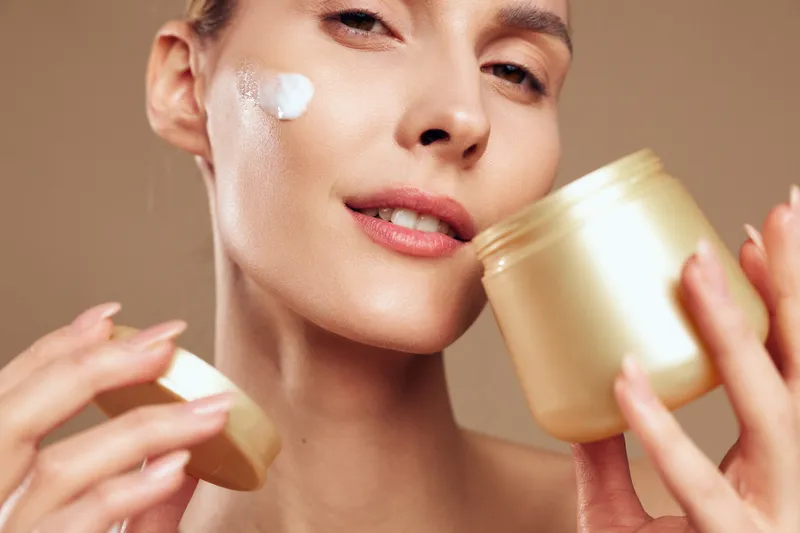 Close up of a woman applying moisturizer from a jar, showcasing smooth skin and a skincare routine, isolated on a soft beige background, casual beauty moment with natural lighting and a soft glow