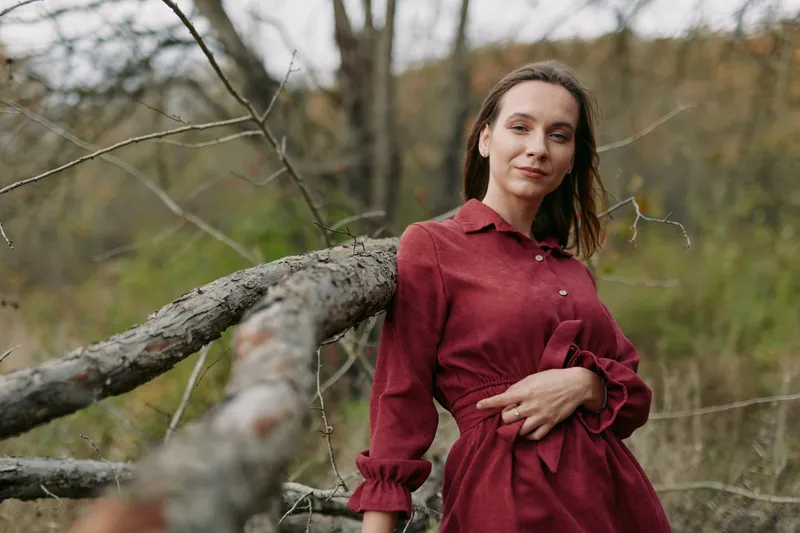 A calm portrait of a woman in a red dress leaning on a fallen tree in a quiet forest, conveying authenticity through natural light, posture, and candid mood.