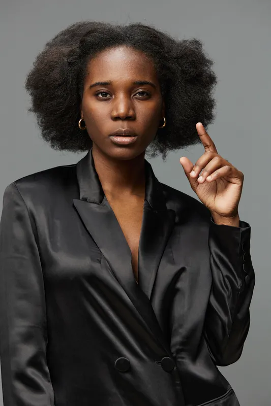 Confident young Black woman in a black satin blazer, pointing with one finger against a soft gray background, embodying empowerment and self assurance