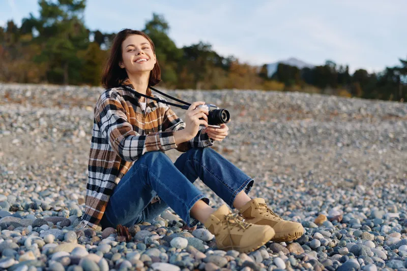 Casual photographer sits on a pebble beach, smiling and holding a camera, relaxed outdoor scene in denim and plaid shirt with natural light and calm sea breeze