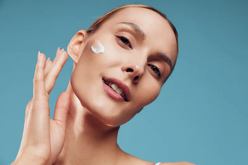 A young woman applies cream to her cheek in a bright studio, showcasing a skincare routine and a moment of self care, emphasizing calm and clean beauty.