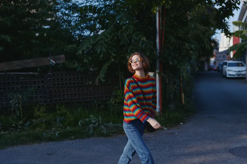 Young adult woman in a colorful striped sweater and denim jeans walking on an urban street, smiling with glasses, carefree lifestyle portrait capturing joyful evening city mood.