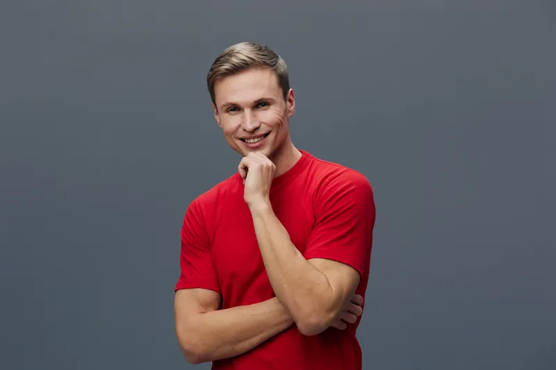 Confident smiling young man with short hair and fair skin wearing casual red t-shirt stands on gray background, looking at camera, cheerful expression, relaxed posture, studio portrait, lifestyle conc
