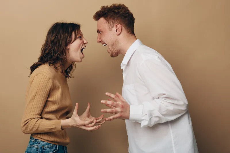 Angry young man and woman shouting at each other in casual clothes on beige background. Concept of conflict, argument, relationship problems, and emotional tension between couple.