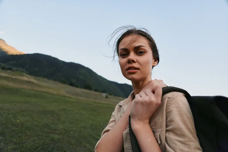 A woman hiking with a backpack stands in a wide meadow, looking toward distant mountains, capturing a moment of travel, nature, and outdoor life.