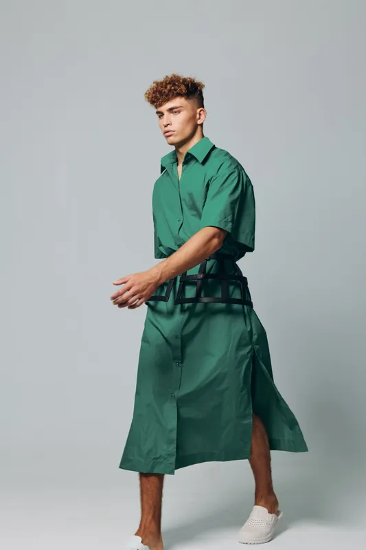 Young man with curly hair wearing green oversized shirt dress and white slip-on shoes posing in studio with neutral background, fashion style and casual look.