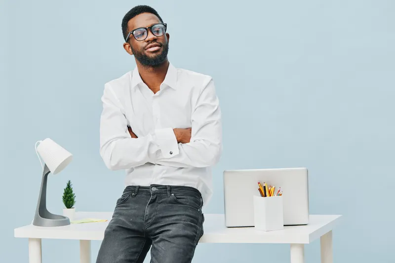 Modern confident man with glasses, casual white shirt, black jeans, standing arms crossed in bright minimalistic office with desk, laptop, and supplies, relaxed professional style, studio shot on ligh