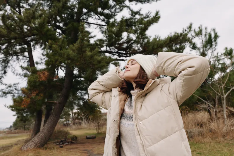 Outdoor woman wearing a beige coat enjoys fresh air outside. She raises her arms and tilts her head back, feeling wind and breeze under a cloudy sky with trees and open field in the background.
