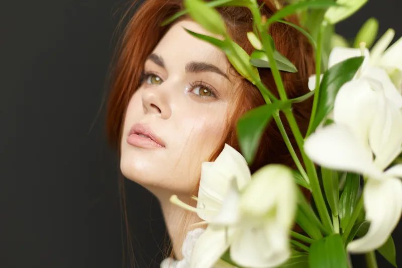 Portrait of a young woman with long red hair surrounded by white lilies, exuding elegance and tranquility in a dark background, perfect for beauty and fashion concepts