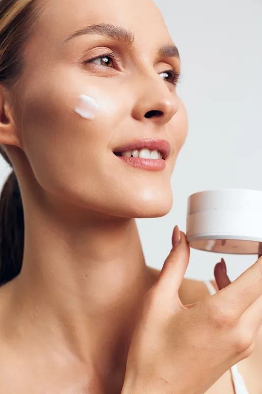 Young woman applying moisturizing cream on cheek, smiling gently, clear skin, closeup portrait, soft natural light, beauty skincare routine, healthy glow