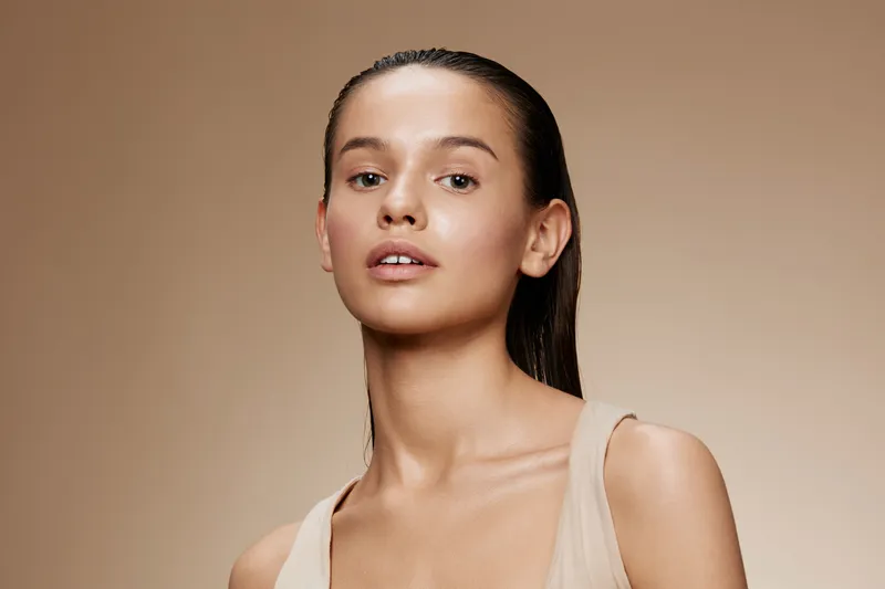 Young female model with a neutral expression, wearing a minimalistic beige outfit against a soft, neutral background, showcasing natural beauty and elegance