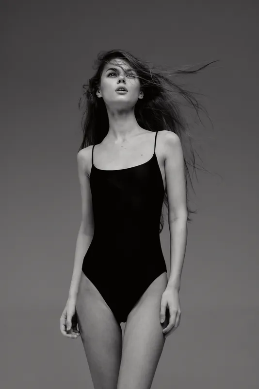 Black and white portrait of a young woman wearing a black swimsuit with windblown hair against a plain background.