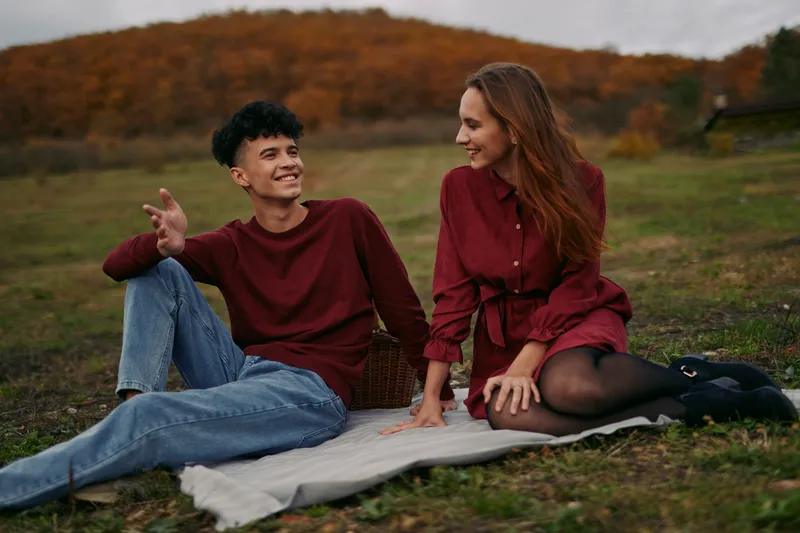 A candid couple sits on a blanket in an open field, sharing a warm smile and an honest moment that feels authentic and credible under soft autumn light.
