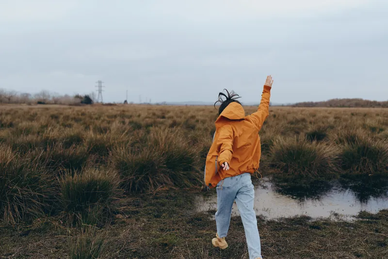 Woman lifestyle outdoors near grassy field, full body walking on path, orange jacket and yellow hat, arm raised toward cloudy sky, peaceful autumn nature scene with wet puddle steps