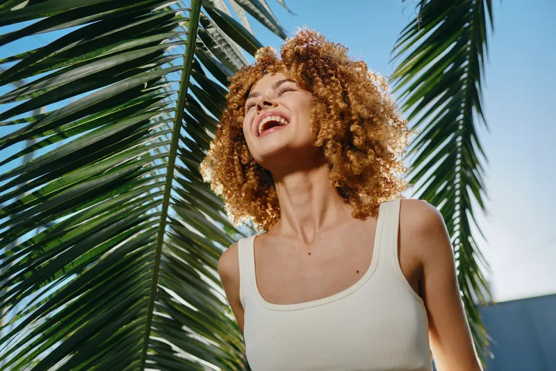 smiling woman with curly hair enjoys sunny tropical outdoors wearing white tank top under clear blue sky with palm leaves background, vibrant summer mood