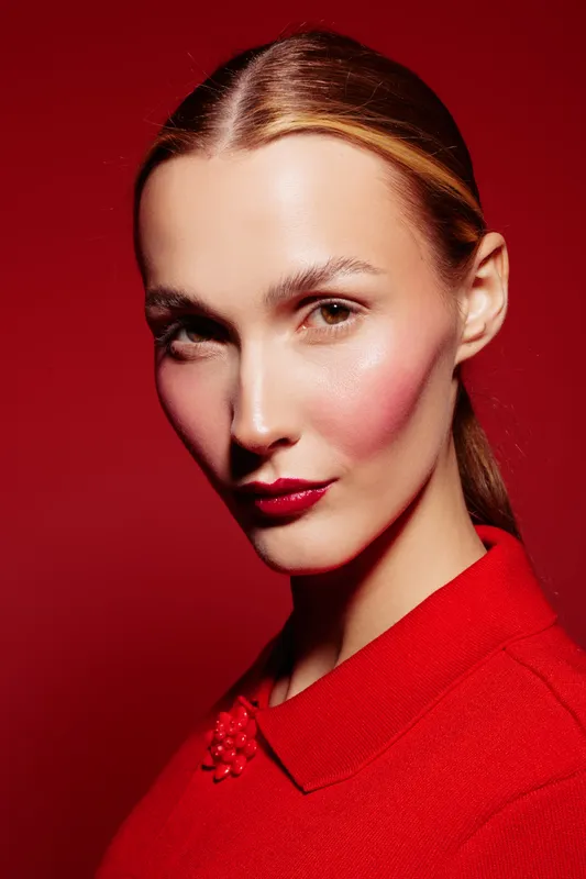Close-up portrait of confident woman with red lipstick and blush wearing red top against red background. Concept of beauty, fashion, makeup, and elegance in studio lighting.