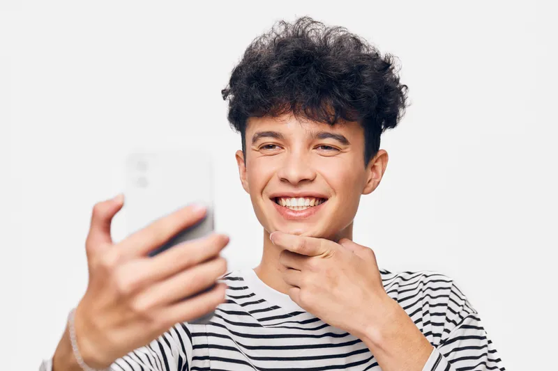 smiling man taking selfie with smartphone, casual striped shirt, curly hair, cheerful expression, bright background, youthful mood
