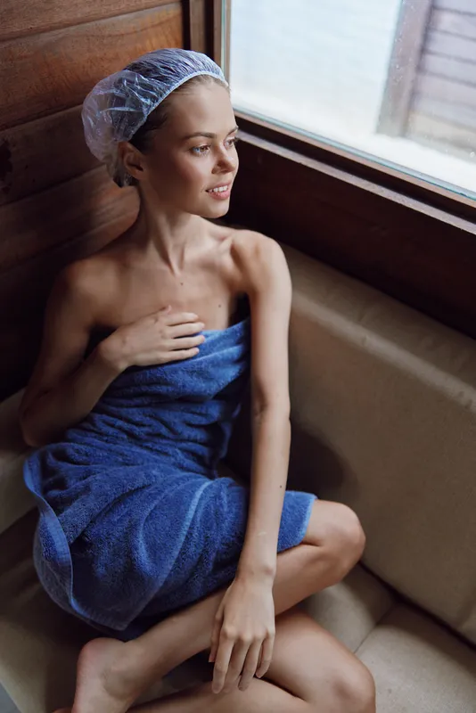 young woman wrapped in blue towel sitting by wooden window, wearing hair cap, relaxed expression, cozy indoor spa setting, natural light bathhouse atmosphere