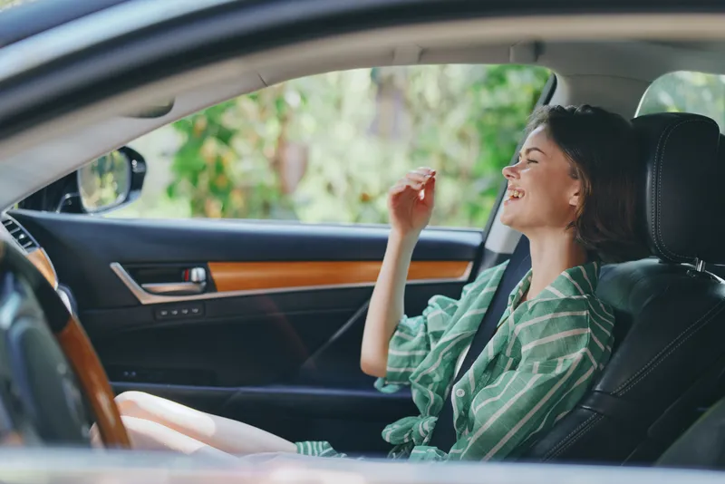 Joyful woman behind the wheel sits in a car seat, smiling while driving, with sunlight streaming through the window and a relaxed vibe.
