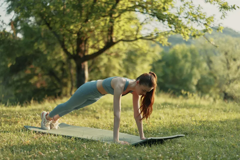 Young athletic woman practicing planks on a mat in a sunny green park, showcasing fitness, determination, and healthy lifestyle surrounded by nature