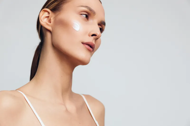 Young woman with clear skin applies moisturizing cream on her cheek, looks away thoughtfully, isolated on light gray background, skincare beauty concept with natural makeup.