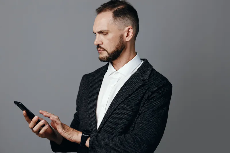 A professional man in a business setting, focused and thoughtful, gestures at a smartphone. An isolated colored background enhances the calm, confident mood, highlighting emotion and concentration.