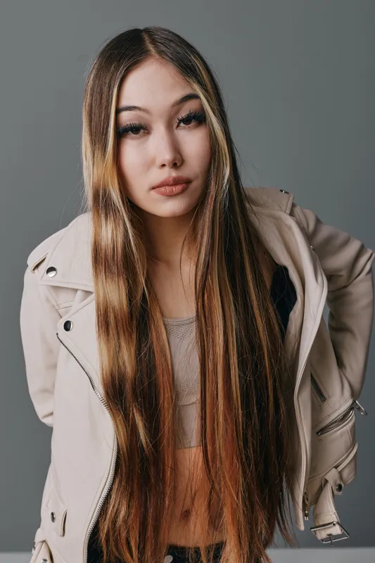 Young woman with long straight hair wearing light jacket stands against gray background looking at camera with neutral expression in studio portrait photo.