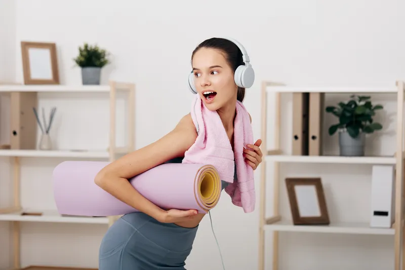Fitness woman with headphones carries a yoga mat and towel in a bright home gym, ready for a focused workout session, blending cardio and yoga elements in a comfortable, private space.