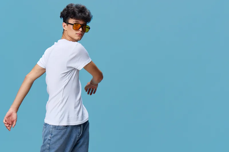 young man wearing white t-shirt and sunglasses posing confidently against blue background with casual style and curly hair in studio setting