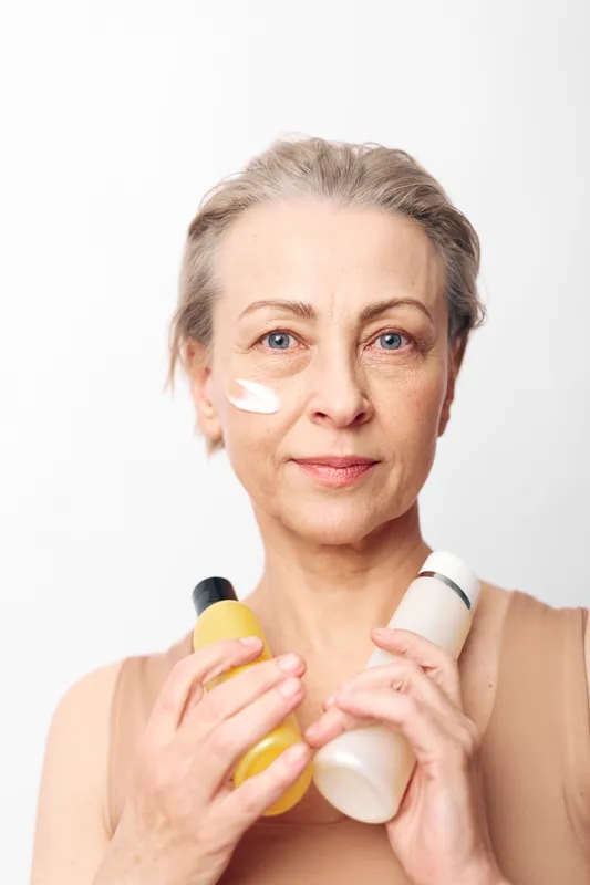 Mature woman with clear skin applies white cream on face holding two cosmetic bottles. Natural skincare concept with facial care products for healthy and radiant skin at home.