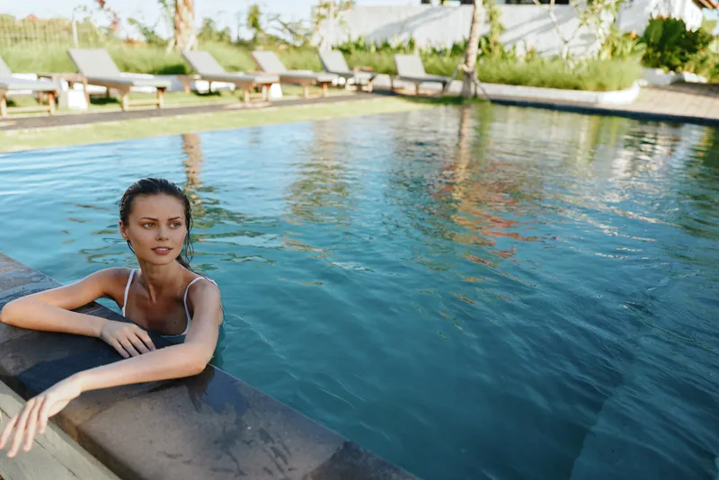 Young woman in swimsuit relaxes in outdoor pool leans on edge looks away enjoys sunny day summer lifestyle vacation leisure activity.