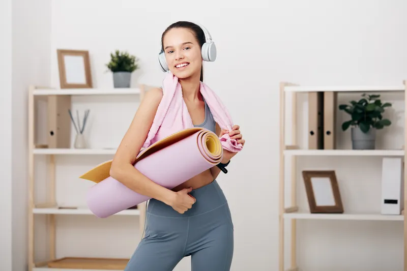 Woman fitness in athletic wear with headphones, carrying a yoga mat and towel in a bright home gym, ready for a workout and sport routine.