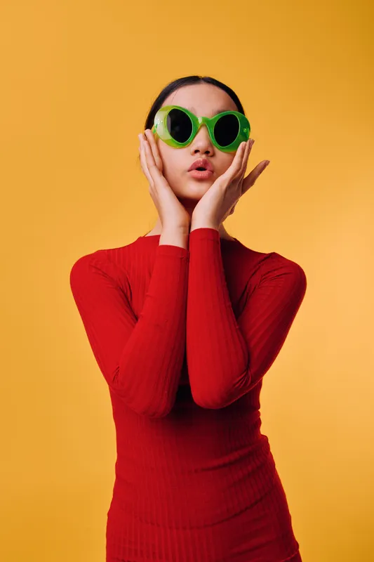 Young woman wearing green round sunglasses and red turtleneck sweater poses with hands on face against yellow background, expressing surprise or curiosity with lips pursed.