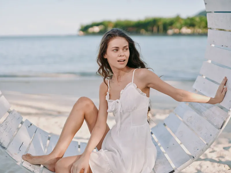A woman in a white sundress sits on a white wooden swing by the beach, gazing to the side as soft waves roll onto the shore and sunlight washes the scene.